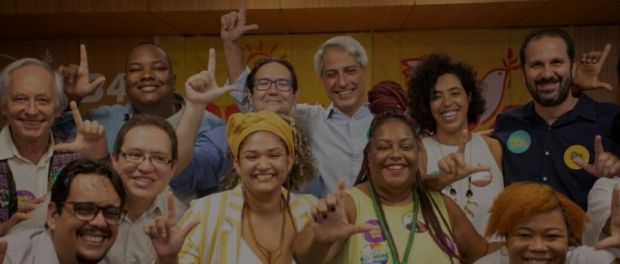 Foto colorida com várias pessoas sem máscara sorrindo e fazendo o L com os dedos. Ao centro está o deputado federal Alessandro Molon, homem, pele clara, com cabelo grisalho e liso, meia idade. Ao seu lado direito está a deputada federal Talíria Petrone, mulher, cabelo cacheado escuro, pele marrom clara.